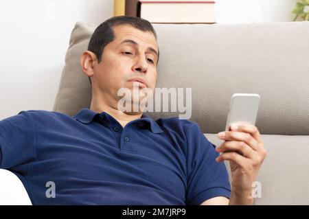 Bored man checking mobile phone sitting on a couch in the living room at home Stock Photo
