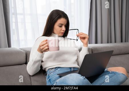 Satisfied woman checks catalogue of new clothes via Notebook holding glasses. Young lady customer rests on soft couch using modern laptop at home, holding cup of tea and glasses in the hands. Stock Photo