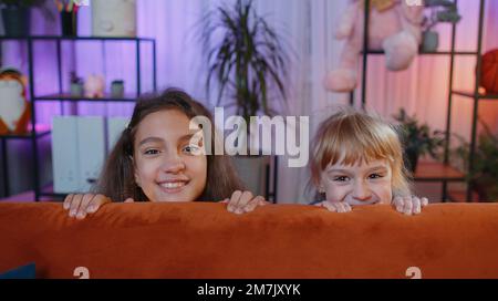 Teenage child and little sister kid girls playing hide and seek peekaboo game. Female smiling siblings children or best friends trying to scare, prank at home play room. Friendship family relationship Stock Photo