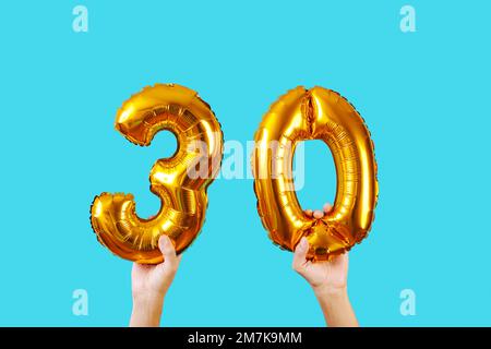 closeup of a man holding two golden number-shaped balloons forming the number 30 on a blue background Stock Photo