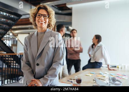 Mature woman in her 40's having a meeting with her team. Happy business woman looking at the camera in a boardroom. Female professional working on a w Stock Photo