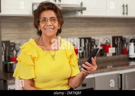 Smiling elderly woman using a smartphone in the comfort of her kitchen. Elderly woman looks and smiles at the camera while holding her phone. Stock Photo