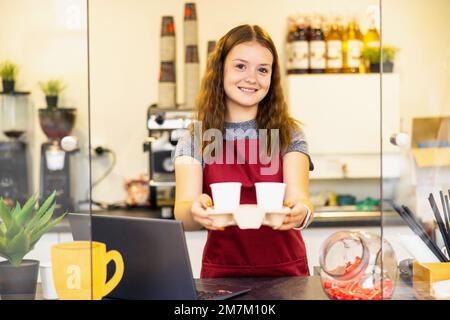 Portrait of a young beautiful woman in red apron, keeps white cup in hand, smiling, looking at camera, on a coffee shop background. Stock Photo