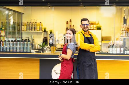 Young baristas in aprons smiling at the camera while standing in front of the bar. Small business concept. Stock Photo
