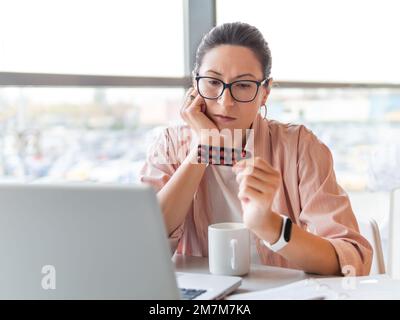 Frowning woman looks on medicine pills while working with laptop. Mental health problems, emotional burnout or headache. Modern office Stock Photo