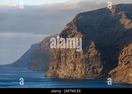 Telephoto view of the sunlit iconic massive cliffs rising out of the calm blue ocean underneath a cloudy sky, viewed from the south Stock Photo