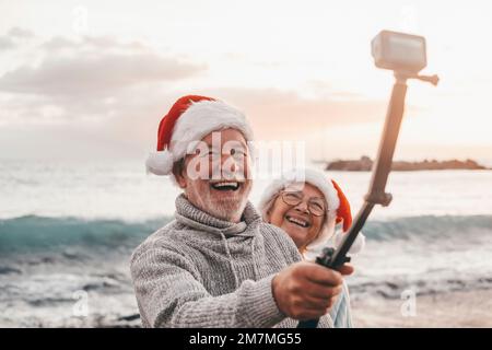 Portrait of two cute old persons having fun and enjoying together at the beach on christmas days at the beach wearing Christmas hats. Looking and holding a camera taking videos of vacations Stock Photo