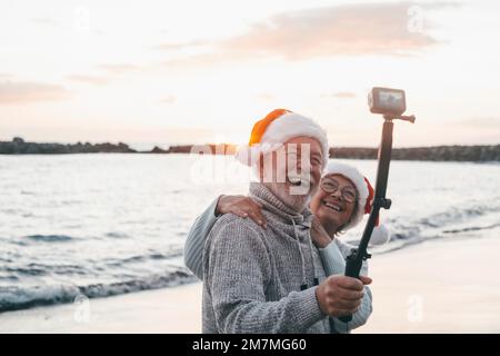 Portrait of two cute old persons having fun and enjoying together at the beach on christmas days at the beach wearing Christmas hats. Looking and holding a camera taking videos of vacations Stock Photo
