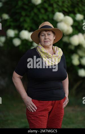 An old woman in fashionable red pants and a stylish scarf poses in the garden with a hat on her head Stock Photo