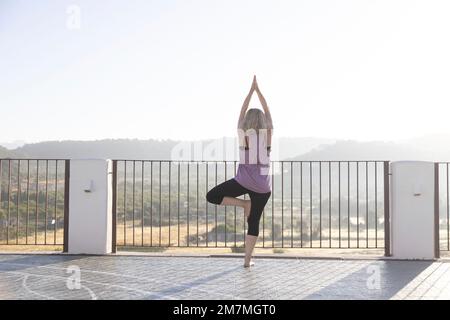 Blonde woman practicing yoga at sunrise on a terrace in Spain Stock Photo