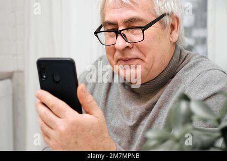 Hoary old man looking at web camera, holding phone, talking with children, wife online. Senior grandpa smiling chatting with friends, family via video Stock Photo