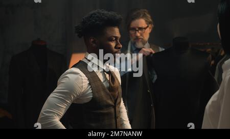 African American young businessman on suit fitting in stylish luxury designer atelier. Tailors put jacket on man. Tailored wedding or business suit. Concept of fashion, handmade and couturier. Stock Photo