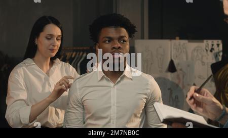 Cinematic shot of tailors taking measurements of African American man for wedding or business suit shirt. Luxury dim atelier or tailoring studio. Concept of fashion, handmade and couturier. Stock Photo