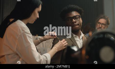 Female tailor measures tailored suit, discusses with client size and fabric. African American client on fitting wedding business jacket in luxury atelier or tailoring workshop. Fashion concept. Stock Photo