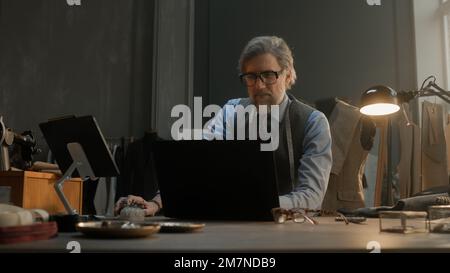 Mature tailor in black framed glasses works on new clothes collection. Man draws modern design of tailorer suit on laptop or tablet computer. Concept of fashion and craftsman job at workshop. Stock Photo
