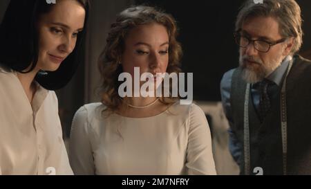 Tailors and woman talking and discussing tailoring beautiful wedding dress. Bride on fitting wedding dress in luxury designer atelier or tailoring studio. Concept of fashion, handmade and couturier. Stock Photo