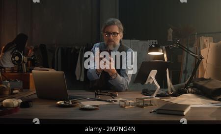 Mature tailor in black framed glasses chatting with client by the phone, taking the order in luxury designer atelier. Woman works with wedding suit on background. Concept of fashion and hand craft. Stock Photo
