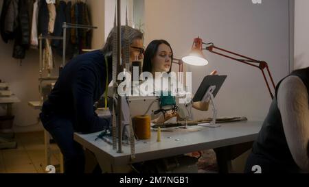Mature tailor with seamstress discussing sketch of suit displayed on tablet. Female dressmaker sits at sewing machine and works with custom apparel. Measure tape on man's neck. Atelier workshop. Stock Photo