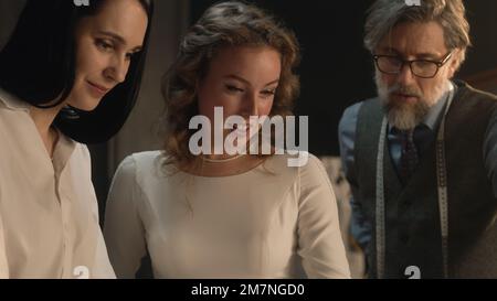 Tailors and woman talking and discussing tailoring beautiful wedding dress. Bride on fitting wedding dress in luxury designer atelier or tailoring studio. Concept of fashion, handmade and couturier. Stock Photo