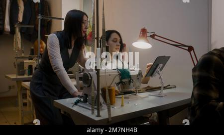 Tailor with seamstress discussing sketch of suit displayed on tablet. Multi ethnic colleagues sitting at sewing machines and tailoring custom apparel. Atelier or sewing workshop. oncept of fashion. Stock Photo