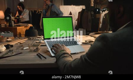 African American fashion designer sits at the table, types on keyboard, takes online orders from customers or creates clothes on laptop with green screen. Atelier view at background. Close up view. Stock Photo