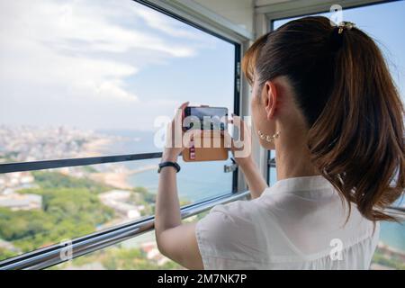 A mature Japanese woman using her mobile phone to take pictures from a cable car cabin of the city and landscape below. Stock Photo