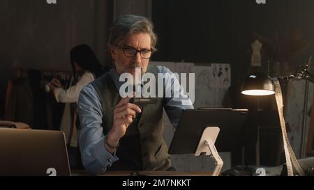 Tailor using tablet, transferring money on bank card, paying for service online during work in luxury atelier. Female tailor works on elegant suit on background. Fashion and entrepreneurship concept. Stock Photo