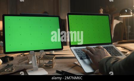 African American fashion designer sits at the table and typing on keyboard, browsing websites or creates clothes on laptop and tablet with green screen. Atelier view at background. Close up view. Stock Photo