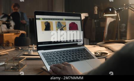 African American client in luxury designer atelier browsing online shop on laptop, watching clothing range, chooses sweatshirts. Concept of fashion and internet shopping. Close up view. Stock Photo
