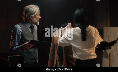 Male and female tailors work on business or wedding suit. Man draws sketch on tablet computer. Mannequin in tailored suit in designer atelier or tailoring studio. Fashion and hand craft concept. Stock Photo