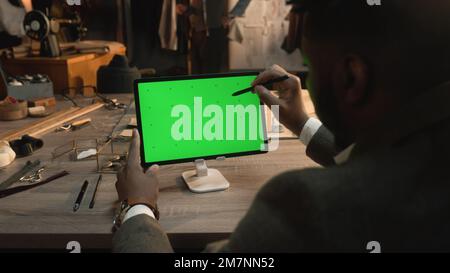 African American tailor sitting at the table in luxury atelier, surfing internet or drawing clothes design on tablet computer with green screen using stylus digital pen. Chromakey. Close-up shot. Stock Photo