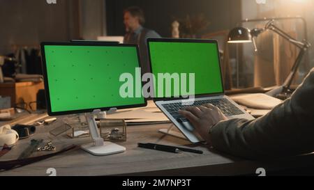 African American fashion designer sits at the table and typing on keyboard, browsing websites or creates clothes on laptop and tablet with green screen. Atelier view at background. Close up view. Stock Photo