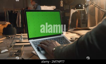African American fashion designer sits at the table, types on keyboard, takes online orders from customers or creates clothes on laptop with green screen. Atelier view at background. Close up view. Stock Photo
