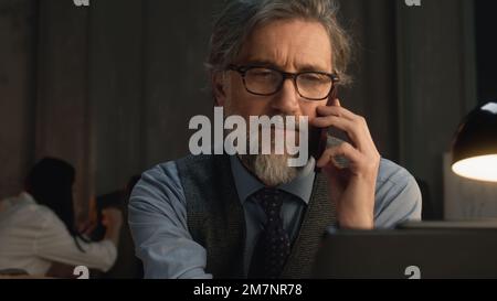 Mature tailor talking by phone and working on design of suit or new clothes collection using digital tablet and laptop. He works in stylish luxury designer atelier. Concept of fashion and handmade. Stock Photo