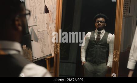 African American man on suit fitting in stylish designer atelier or in dim luxury clothing shop. Tailor puts jacket on man in front of the mirror. Tailored wedding or business suit. Fashion concept. Stock Photo