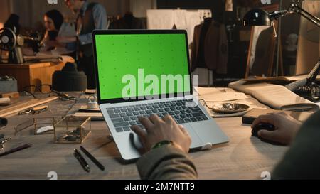 African American fashion designer sits at the table, types on keyboard, takes online orders from customers or creates clothes on laptop with green screen. Atelier view at background. Close up view. Stock Photo