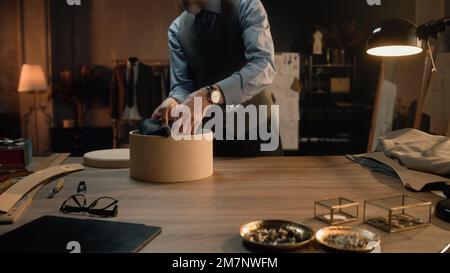 Male mature tailor works in luxury designer atelier or tailoring studio. He packs tailored product for client in box. Tailored suits on background, table with tools. Fashion and hand craft concept. Stock Photo