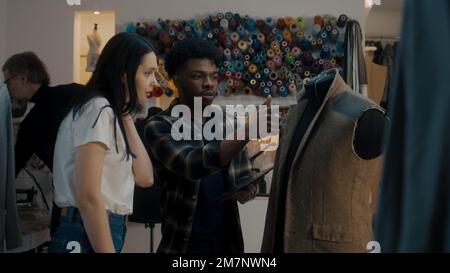 African American fashion designer talks with seamstress in atelier and shows sketch of future suit on digital tablet computer. Mannequin in unfinished tailored suit. Concept of fashion and hand craft. Stock Photo