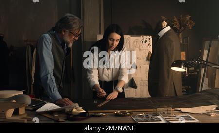 Male and female tailors working on sketch pattern of business or wedding suit. Mannequin with tailored shirt and jacket in stylish luxury designer atelier or tailoring dim studio. Fashion concept. Stock Photo