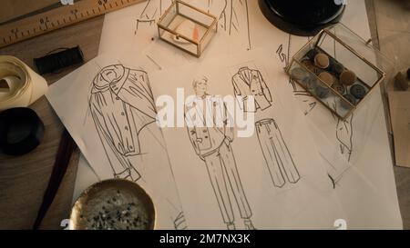 Cinematic shot of table with drawings, needles, threads, buttons, ribbons, soap and ruler. Sketches of clothes, suits and jackets. Designer atelier or tailoring studio. Fashion and hand craft concept. Stock Photo