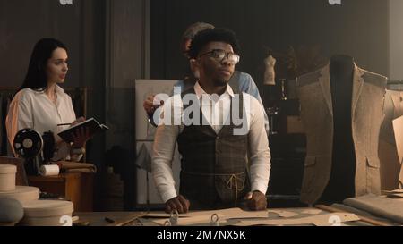 Male tailor measures African American client, discusses with female seamstress design of tailored suit. Woman making notes and writes down the measurements. Concept of fashion and hand craft. Stock Photo