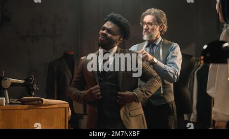 African American man on suit fitting in stylish luxury designer atelier or tailoring studio. Tailor puts jacket on man. Tailored wedding or business suit. Concept of fashion, handmade and couturier. Stock Photo