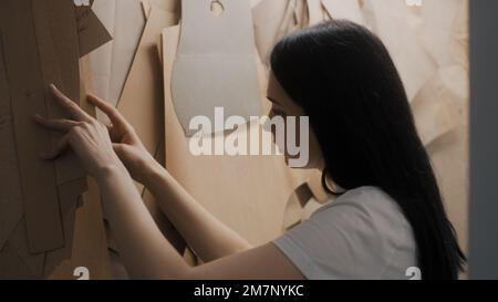 Female dressmaker chooses pattern for tailoring new clothes collection in studio. Seamstress takes patterns from rack while working on custom apparel. Atelier or sewing workshop. Concept of fashion. Stock Photo
