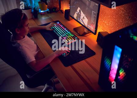 Image of immersed teenage gamer boy playing video games on computer in dark room wearing headphones and using backlit colorful keyboard Stock Photo