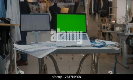 Dolly shot of laptop with green screen and digital tablet computer with sketch on table in atelier workshop. Tailor and seamstress work on background. Concept of fashion and technologies in business. Stock Photo