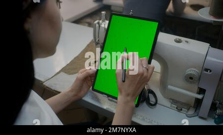 Wear designer or seamstress sits at sewing machine, watches at tablet, keeps stylus in hand and touches screen. Green screen displayed on gadget. Atelier workshop. Concept of fashion. Close up shot. Stock Photo
