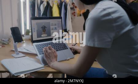 Female fashion designer draws future clothes in 3D modeling software on laptop using digital tablet computer and stylus. She works in atelier workshop. Concept of fashion and technologies in business. Stock Photo