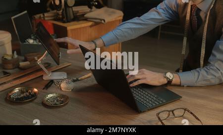 Mature tailor chooses materials and colors of textile, works on new clothes collection. Man draws modern design of tailored suit on laptop or tablet. Concept of fashion and craftsman job at workshop. Stock Photo