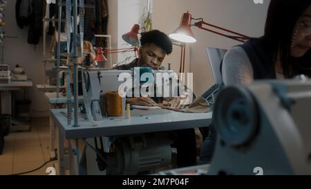 African American male seamstress sits at sewing machine and tailoring wedding suit by table with laptop. Multi ethnic colleagues working on custom apparel at background. Atelier or sewing workshop. Stock Photo