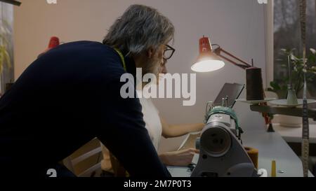 Mature tailor with seamstress discussing sketch of suit displayed on tablet. Female dressmaker sits at sewing machine and tailoring custom apparel. Colleagues working at background. Atelier workshop. Stock Photo